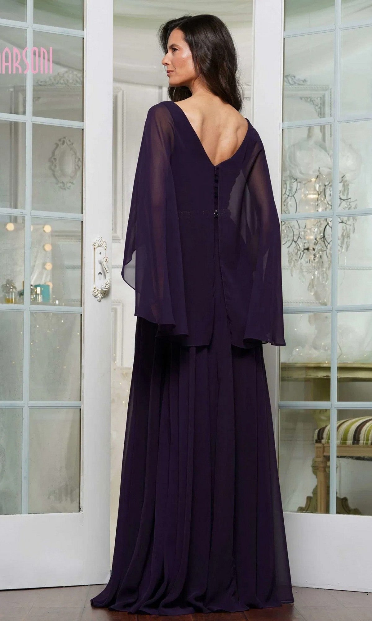 A woman stands facing glass doors in the Marsoni MV1350 long formal dress, featuring dark purple chiffon, sheer sleeves, button-back detailing, an embellished waistbelt, and a V-shaped back neckline.